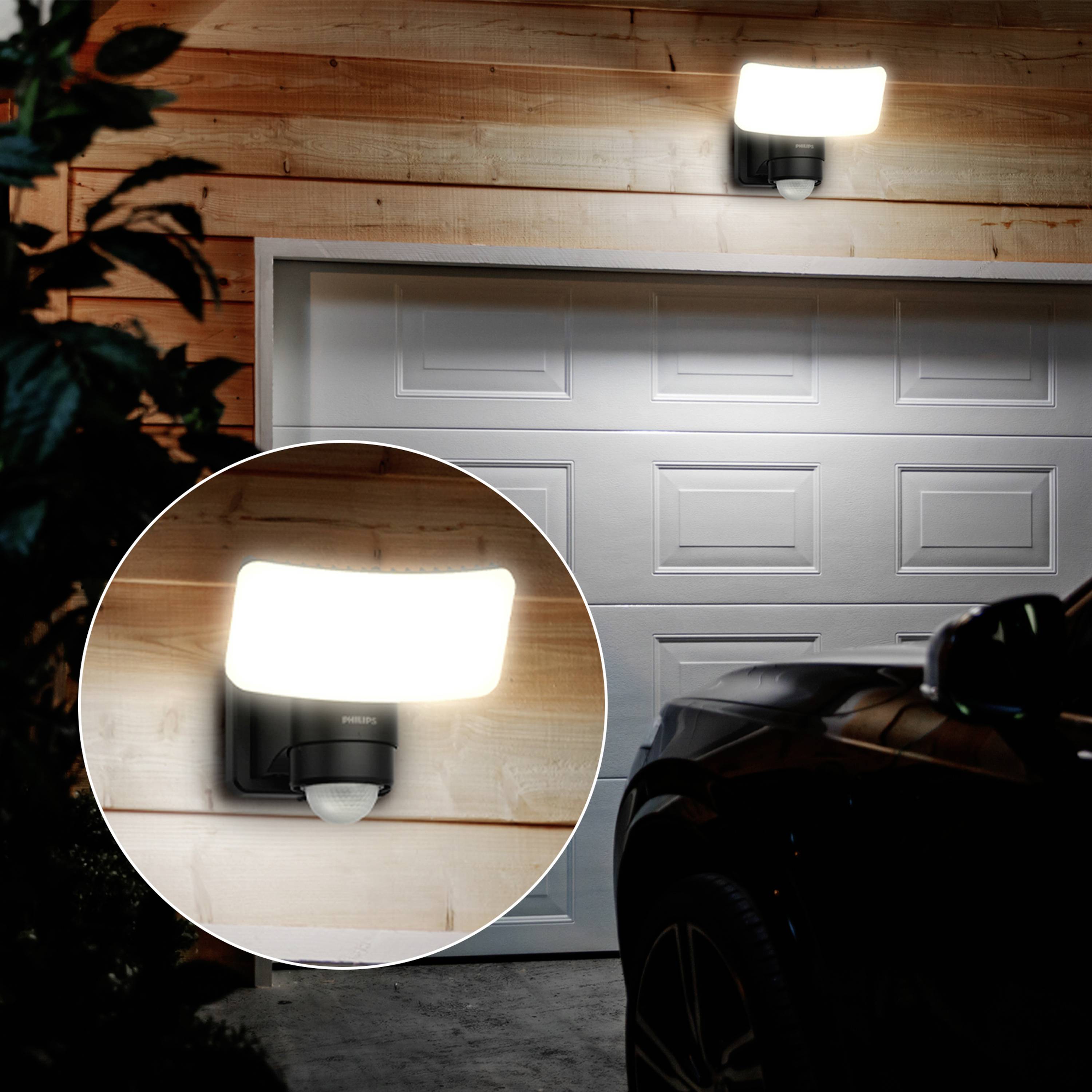 An exterior wall light illuminates in the darkness above a closed garage door, accentuated by wooden cladding. A vehicle is positioned in the foreground.