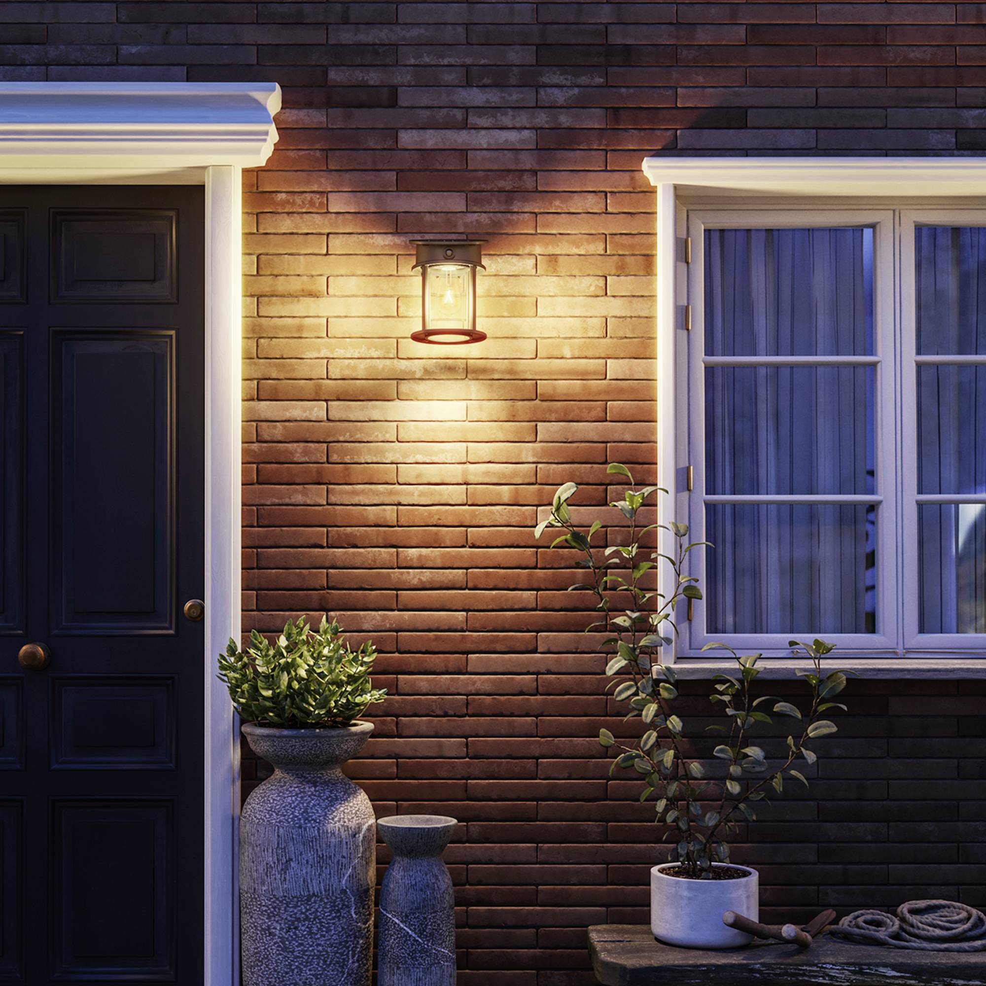 A front door and a window on a brick facade, illuminated by a warm lamp; beside it, plants in pots.