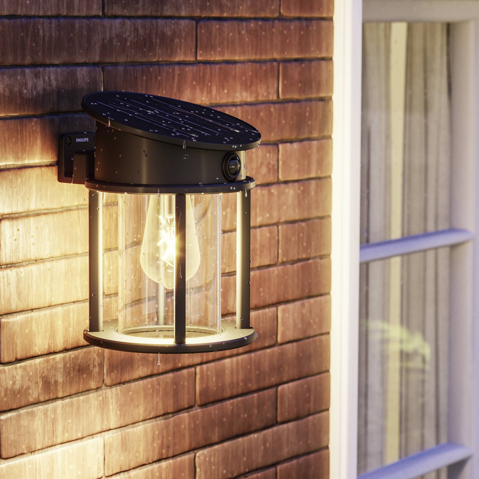 Exterior lamp on a brick wall, illuminated at dusk, beside a window. The lamp has a classic, cylindrical shape.