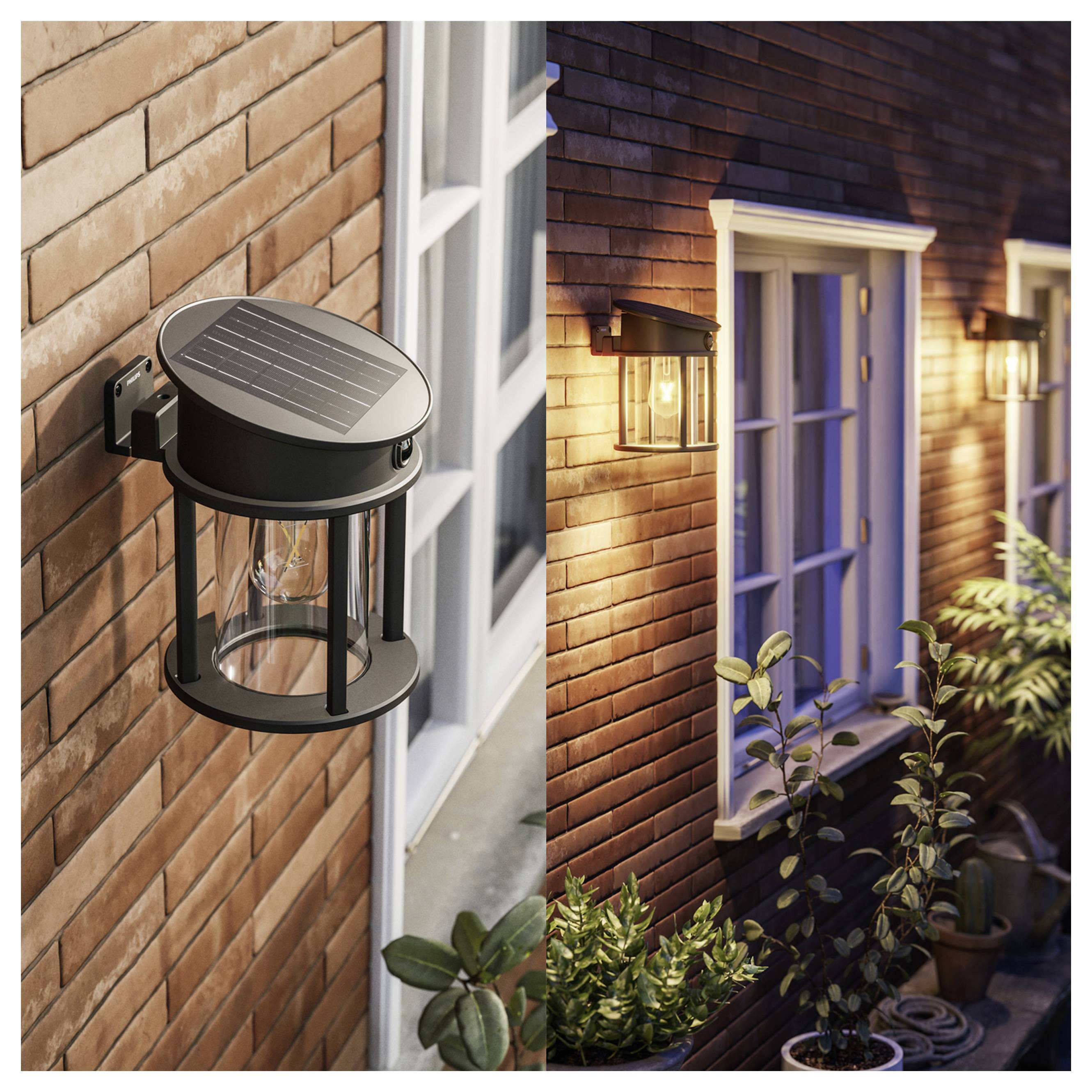Outdoor wall-mounted solar lantern on a brick house exterior, shown lit beside a window and door at dusk with nearby plants.<br>