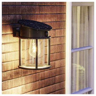 Black outdoor wall lantern with a glowing bulb mounted on a shingled house wall beside a window.<br>