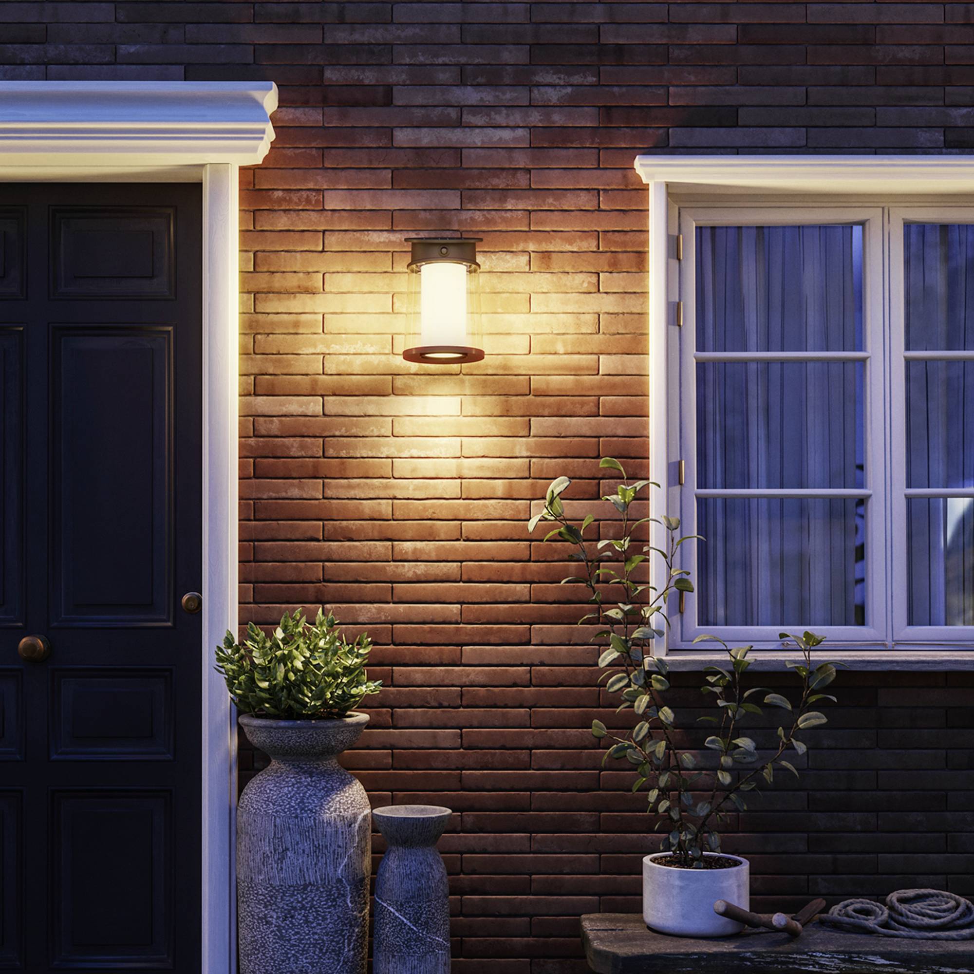 Red brick wall with an illuminated exterior lantern, a black door and a window. Potted plants decorate the entrance.