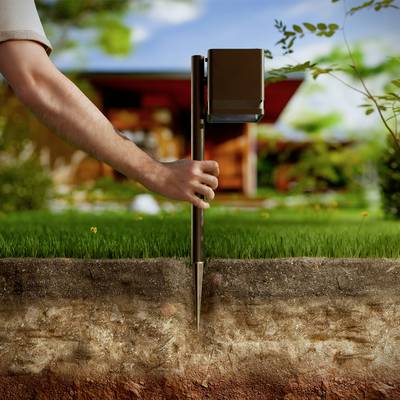 A hand holds a soil testing device inserted into the ground; behind it, a modern house with landscaped garden.