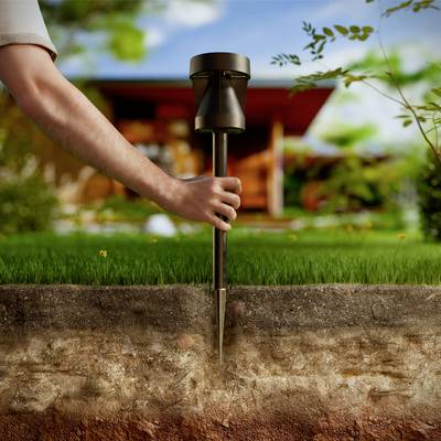 Close-up of a hand inserting a soil moisture metre into the ground of a lawn, with a blurred house in the background.