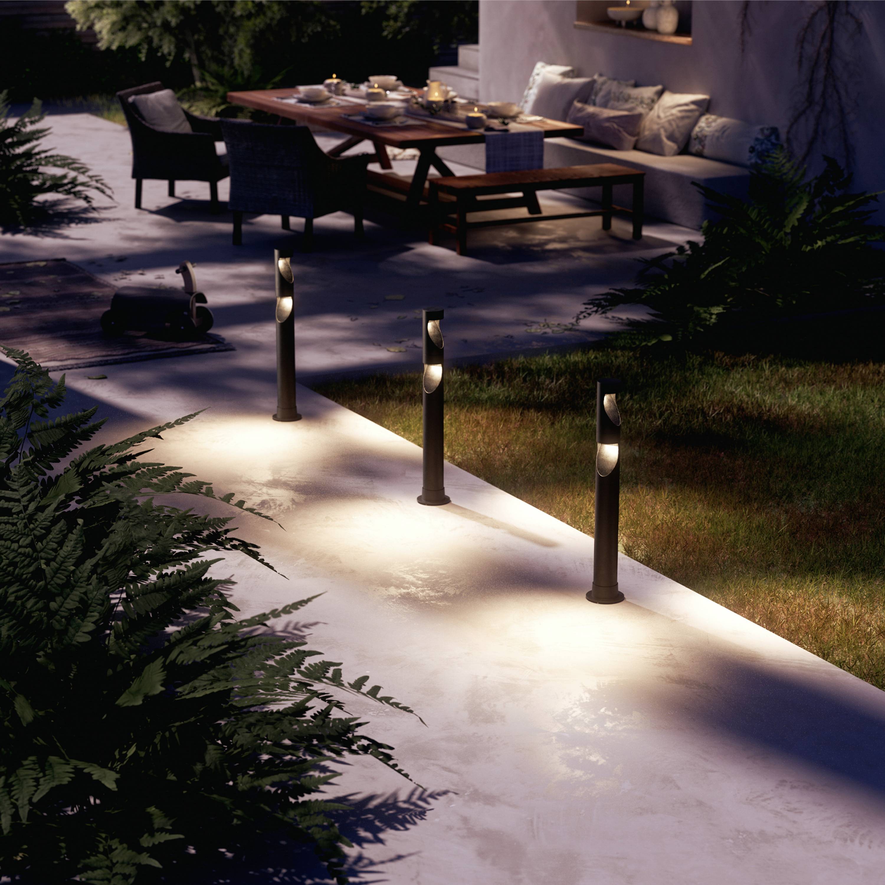 A garden path with several modern lamps illuminating the way. In the background, a table and chairs are positioned on a terrace.