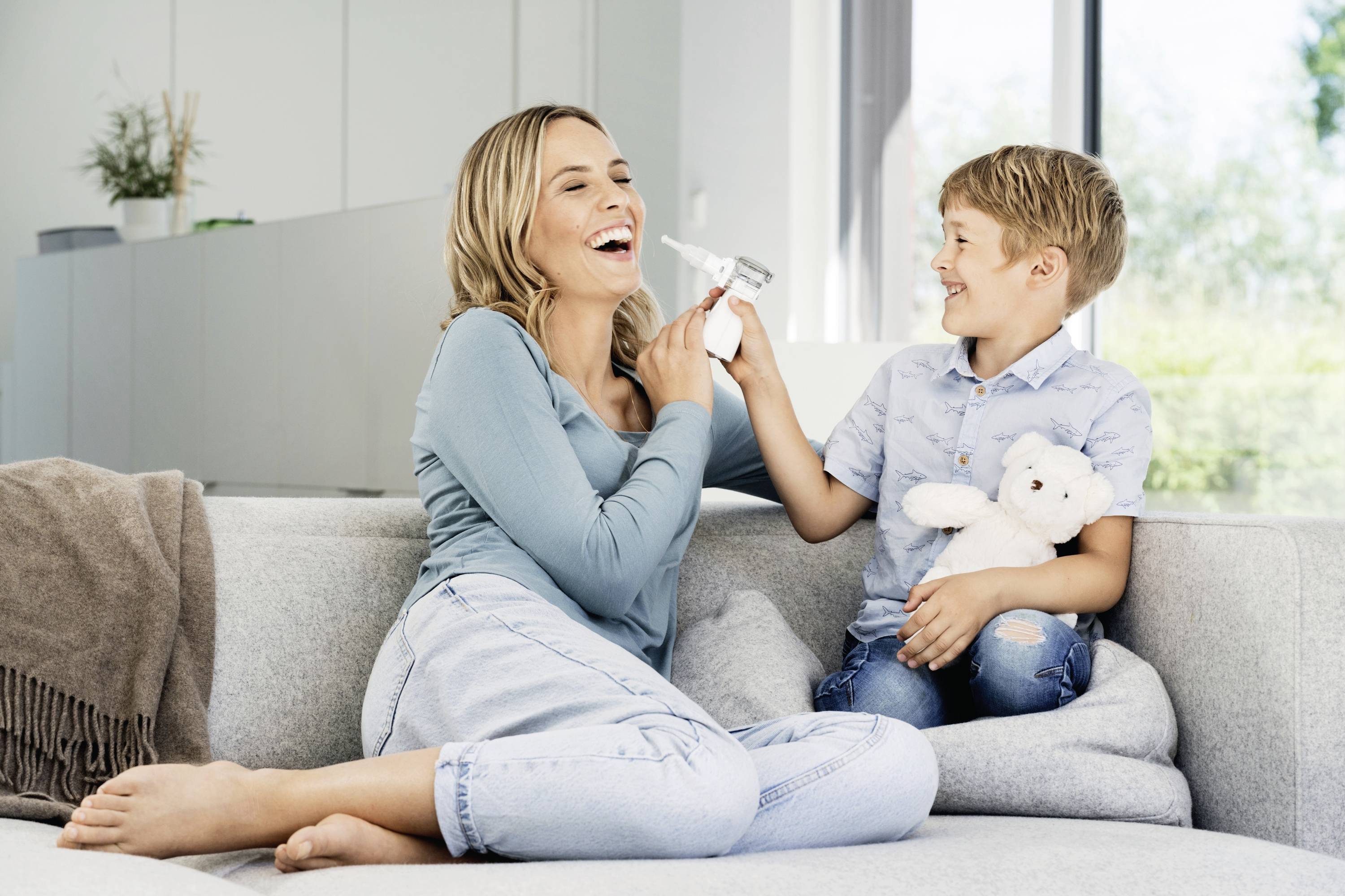 A laughing woman sits on a sofa, while a child sits beside her with a soft toy and shows her a razor blade.