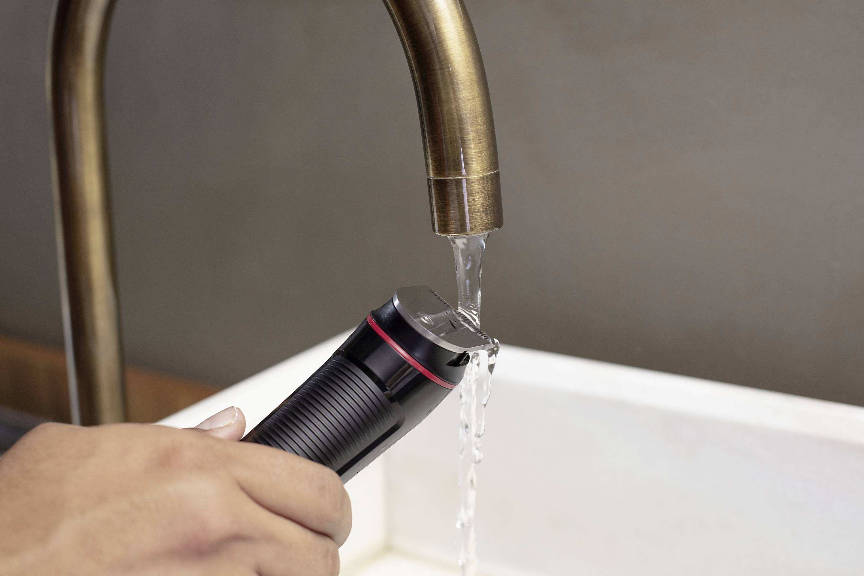 A hand holds a black electric razor under a running tap over a white sink.