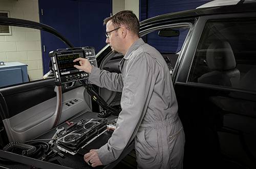 A mechanic in a grey overall is operating a diagnostic device inside a car to check the vehicle's status.