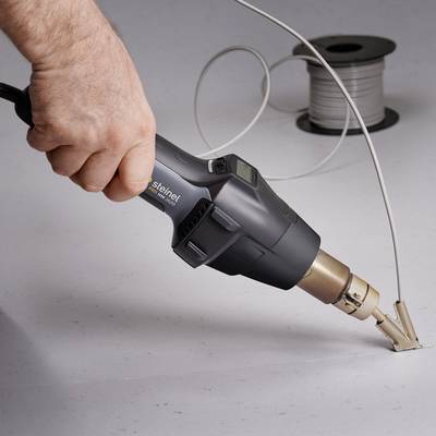 A person is holding a hairdryer and directing it towards electrical cables on a work surface. A cable reel is lying in the background.