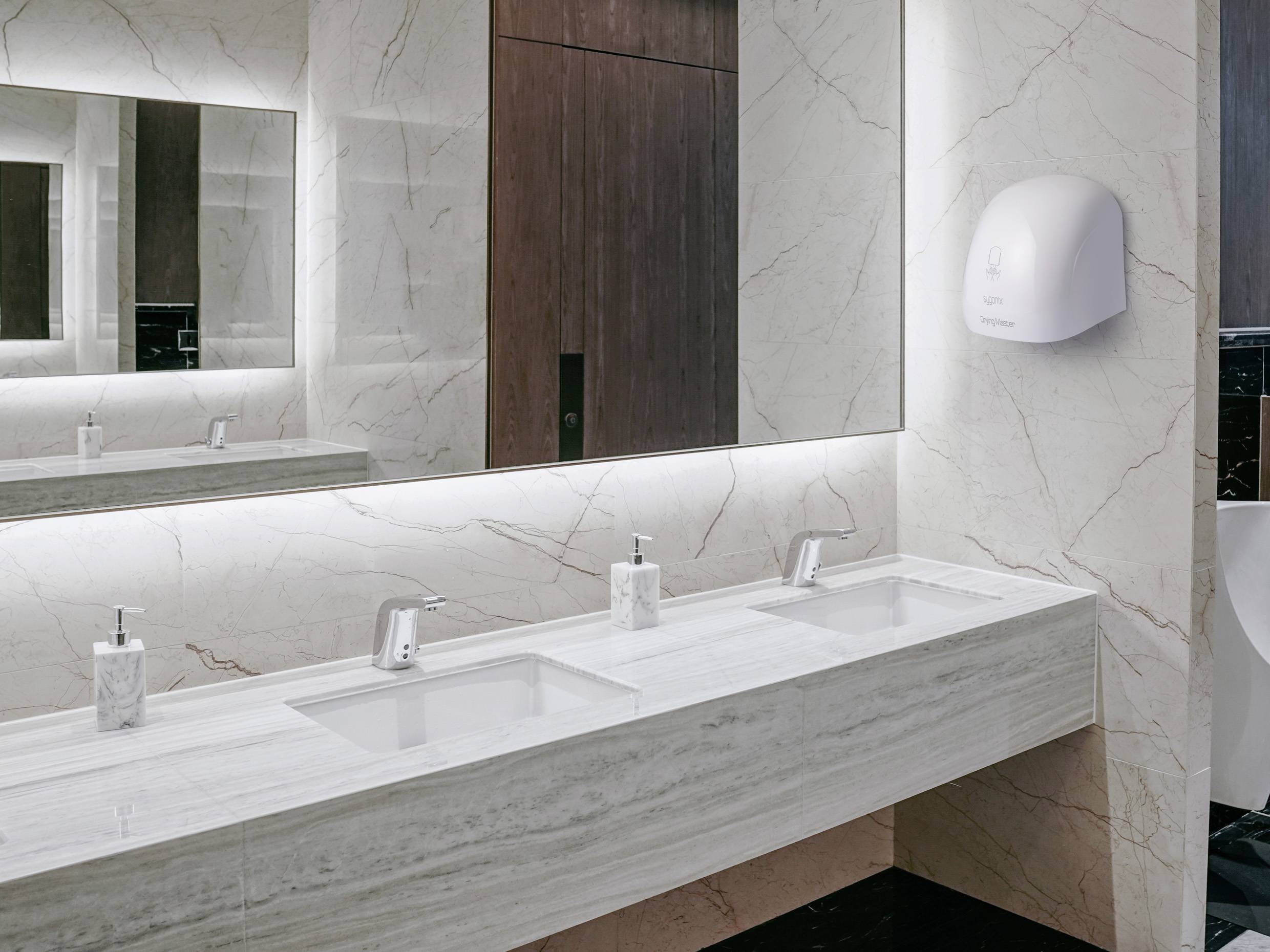 Modern bathroom with marble sinks, mirrors, and a wall-mounted hand dryer. Soft lighting creates an elegant ambiance.