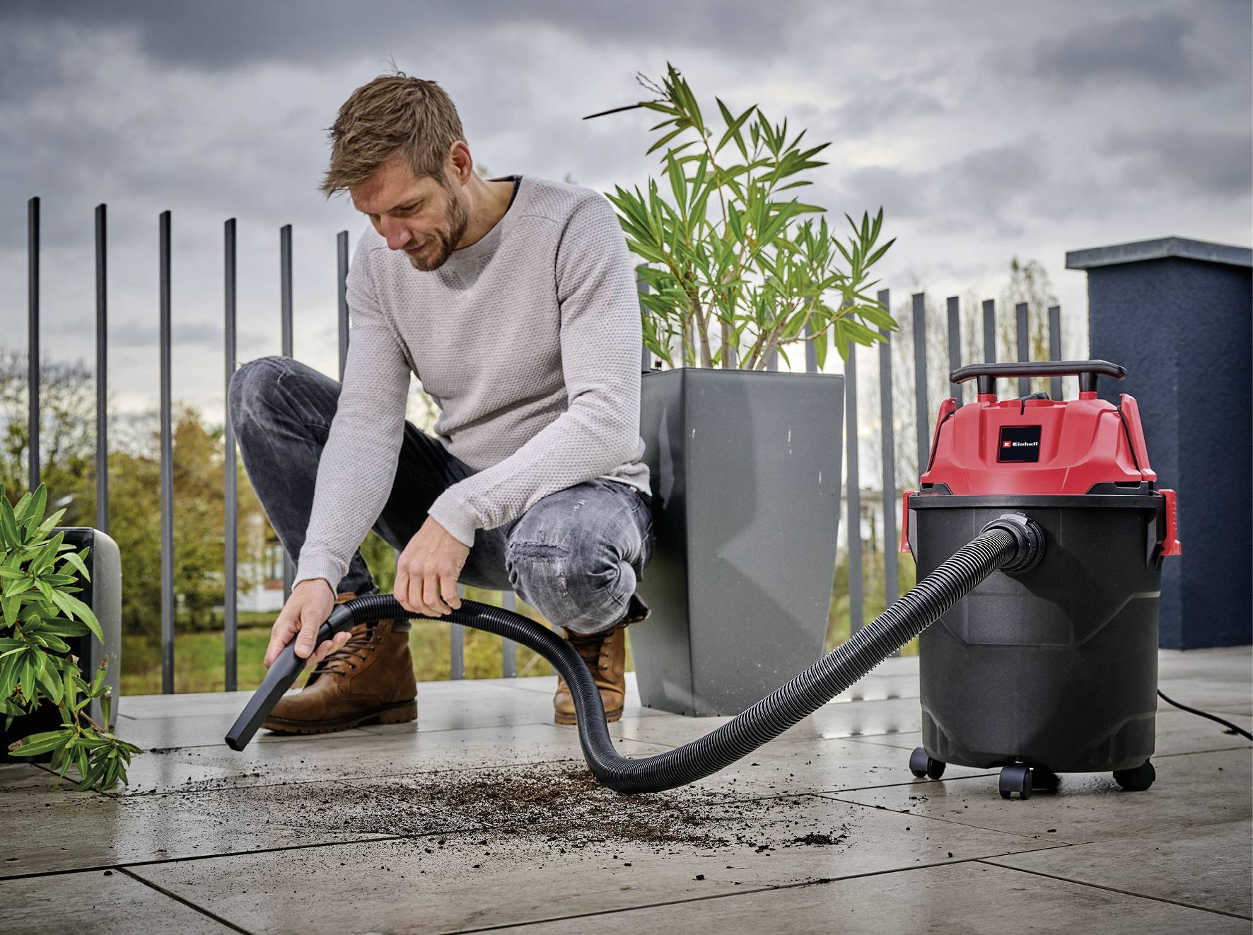 A man is cleaning earth from a patio using a wet and dry vacuum cleaner. Plants and a railing are visible in the background.
