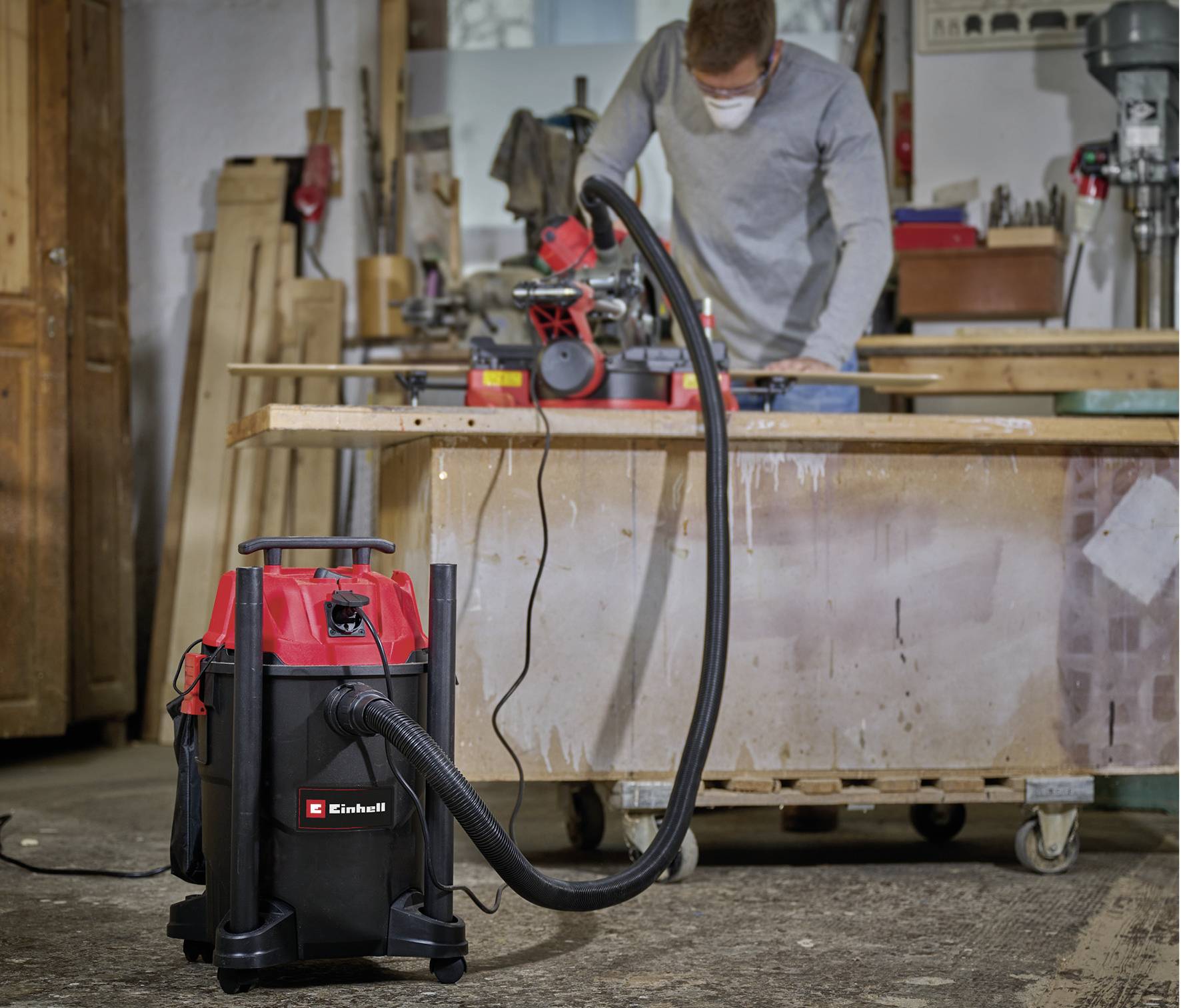 A person is working at a table in a workshop using an electric grinding tool. An industrial vacuum cleaner stands in the foreground.