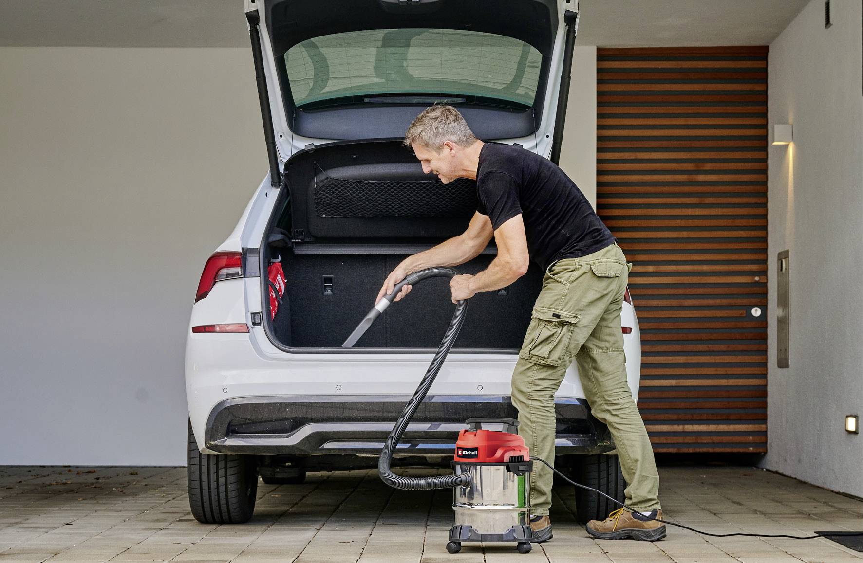 A man is cleaning the boot of a white car with a vacuum cleaner. The car is parked in a garage with wooden walls.