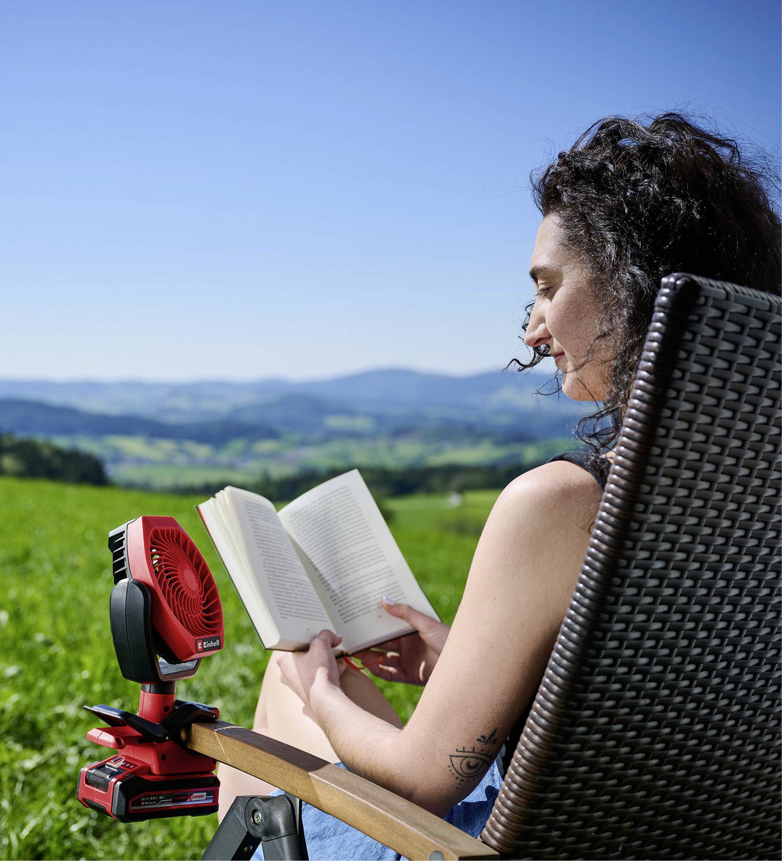 A woman is sitting on a chair outdoors reading a book. Next to her is a small red fan. In the background is a green landscape.