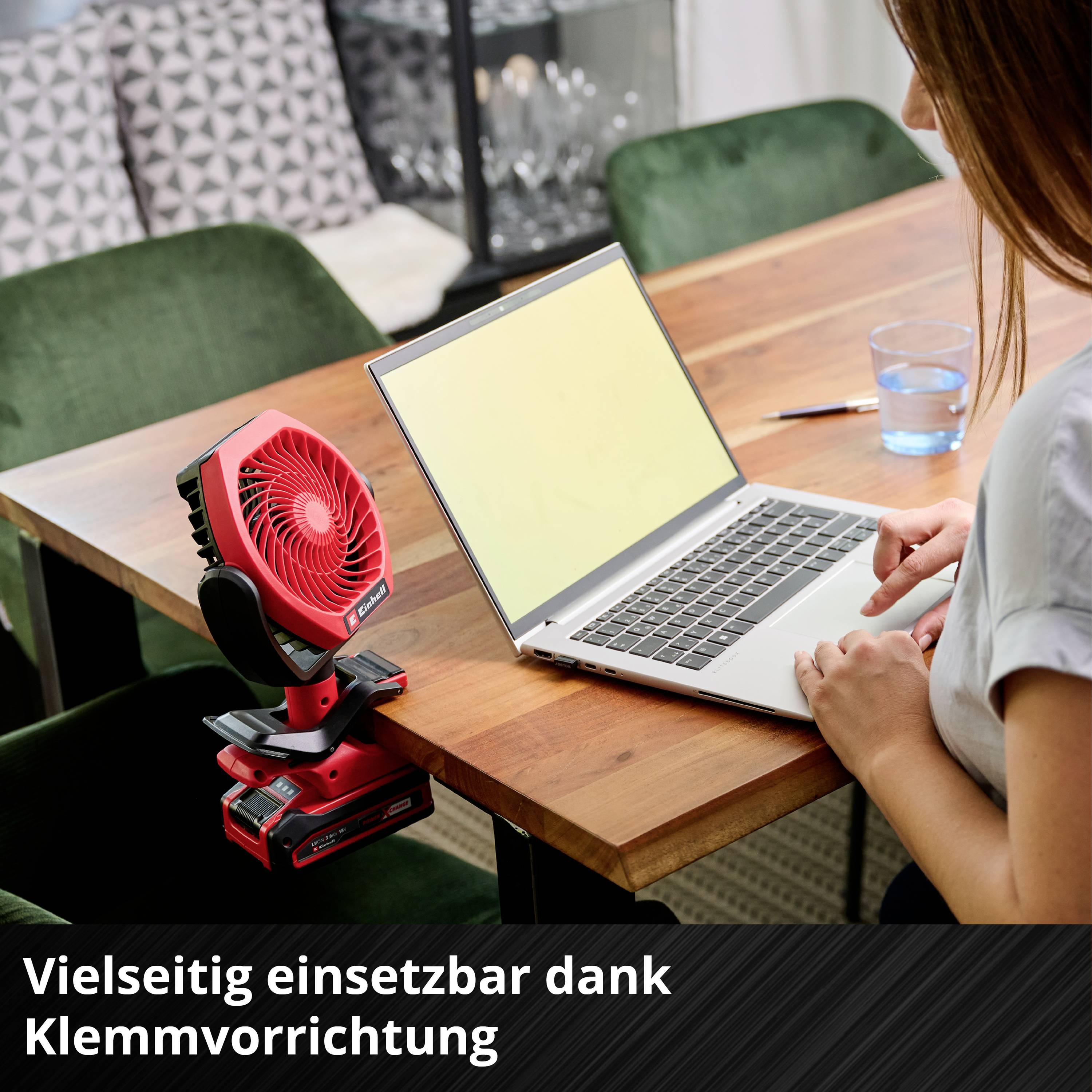 Laptop on a wooden table next to a small red fan, on the left, in front of a green armchair. Text below: 'Versatile thanks to its clip attachment'.