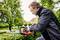 A man is trimming a hedge with an electric hedge trimmer in a garden; he is wearing safety glasses and ear plugs, surrounded by trees and residential houses.