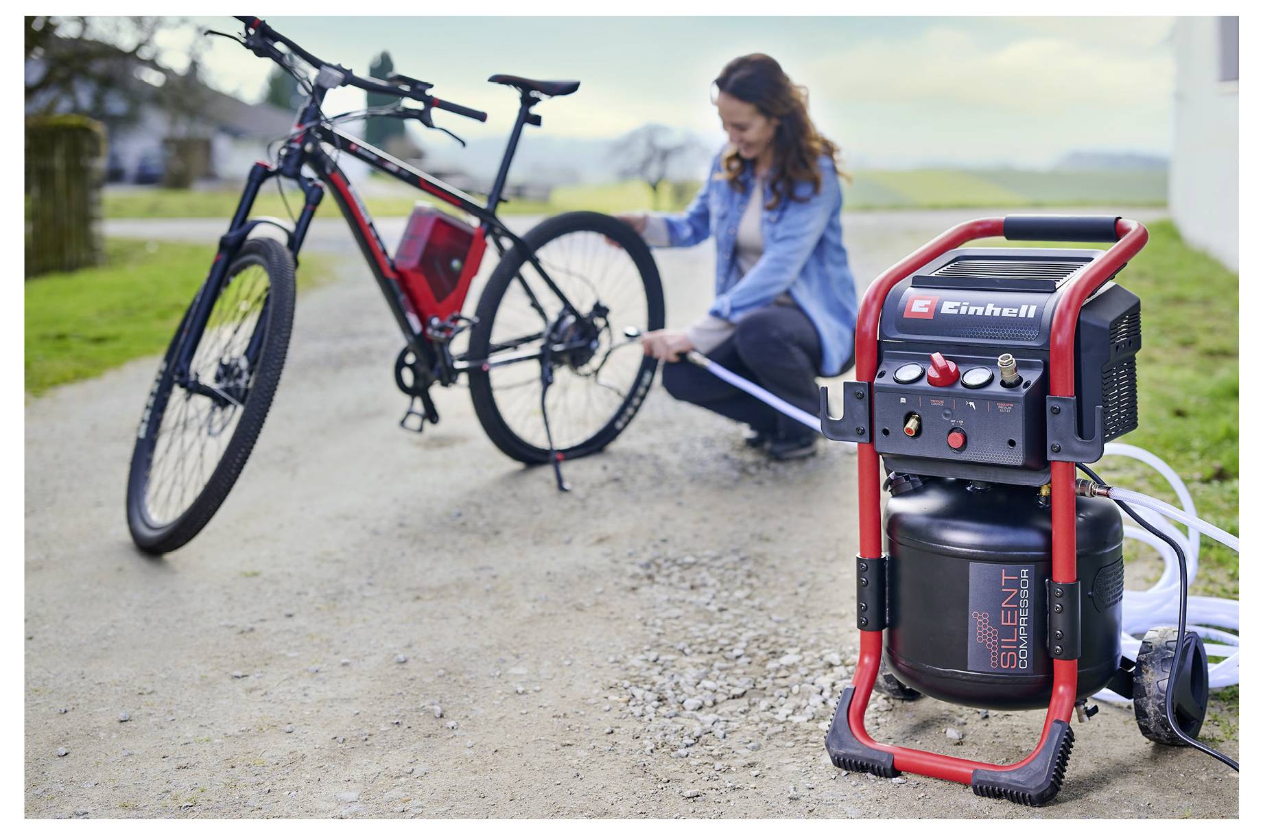 Woman inflating a bicycle tire with a portable Einhell air compressor beside a bike on a roadside.