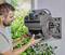 A man is installing an automatic hose reel on a wall, surrounded by green vegetation.