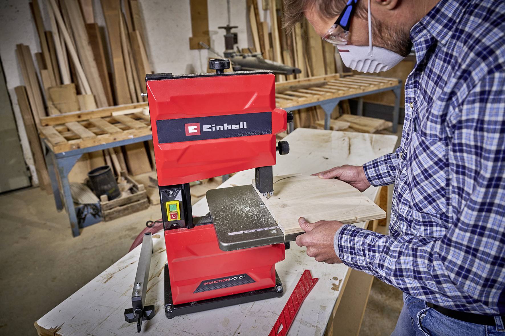 A person in a workshop is using a red bandsaw to cut a piece of wood. They are wearing safety glasses and a mask.