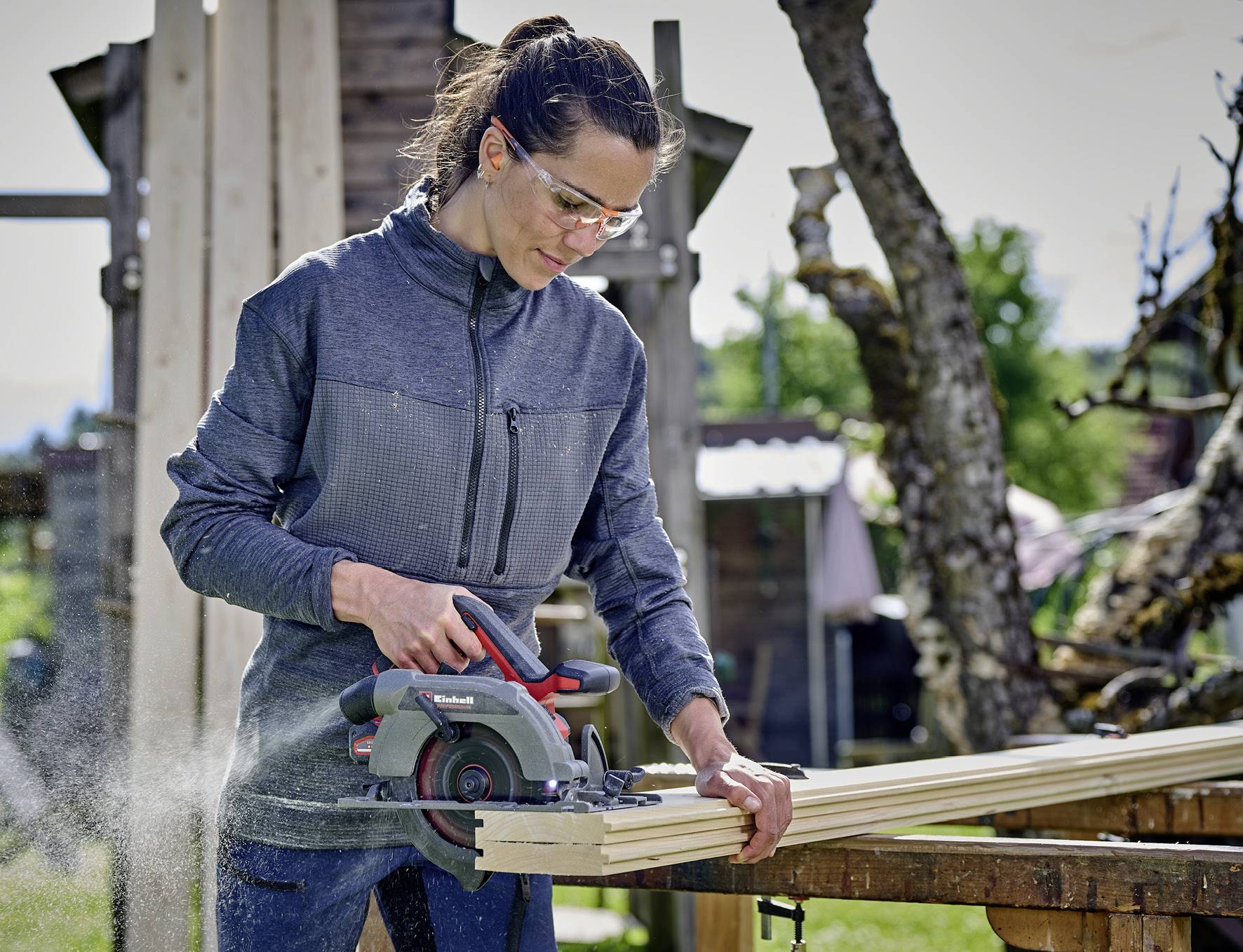 A woman wearing safety goggles is working outdoors with a circular handsaw to cut wooden battens.