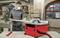 A man is working at a circular table saw in a workshop, wearing a protective mask and cutting wood. Machines and shelves are visible in the background.