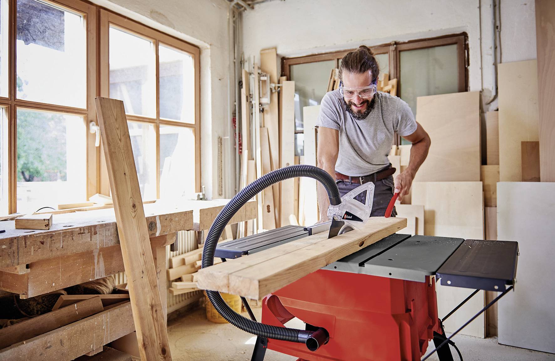 A person uses a table saw to cut wooden planks in a workshop filled with carpentry tools and materials.