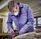 A man wearing safety glasses and a mask is working in a workshop, using an electric tool on a piece of wood.