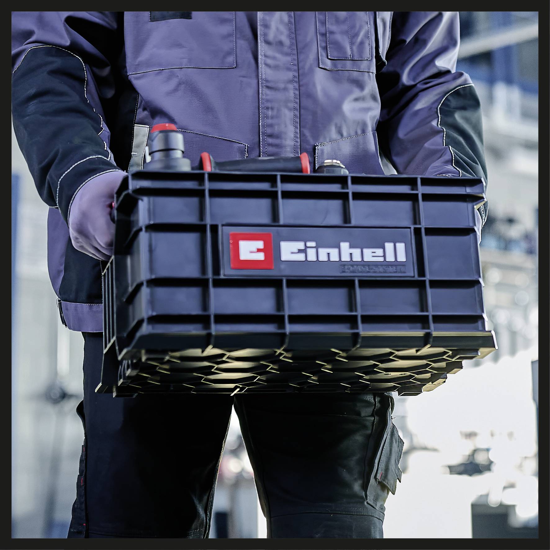 A person in workwear is holding a black toolbox with the 'Einhell' logo. The background shows a blurred industrial landscape.