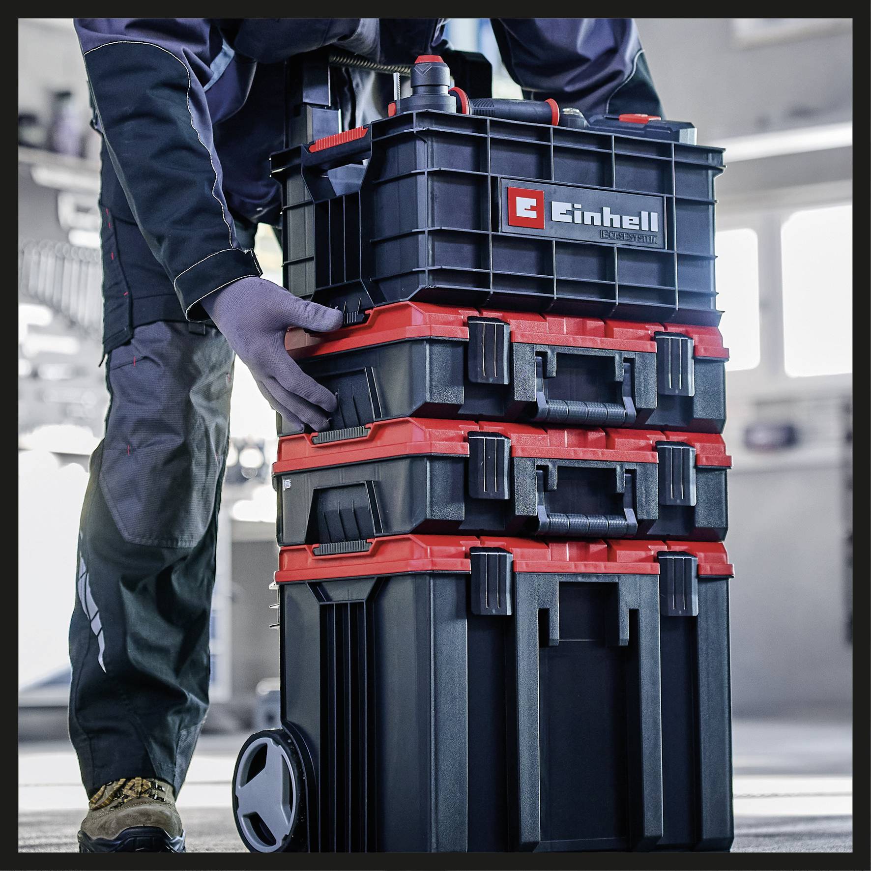 A person is stacking several black tool cases with red locks and a red logo in a workshop environment.