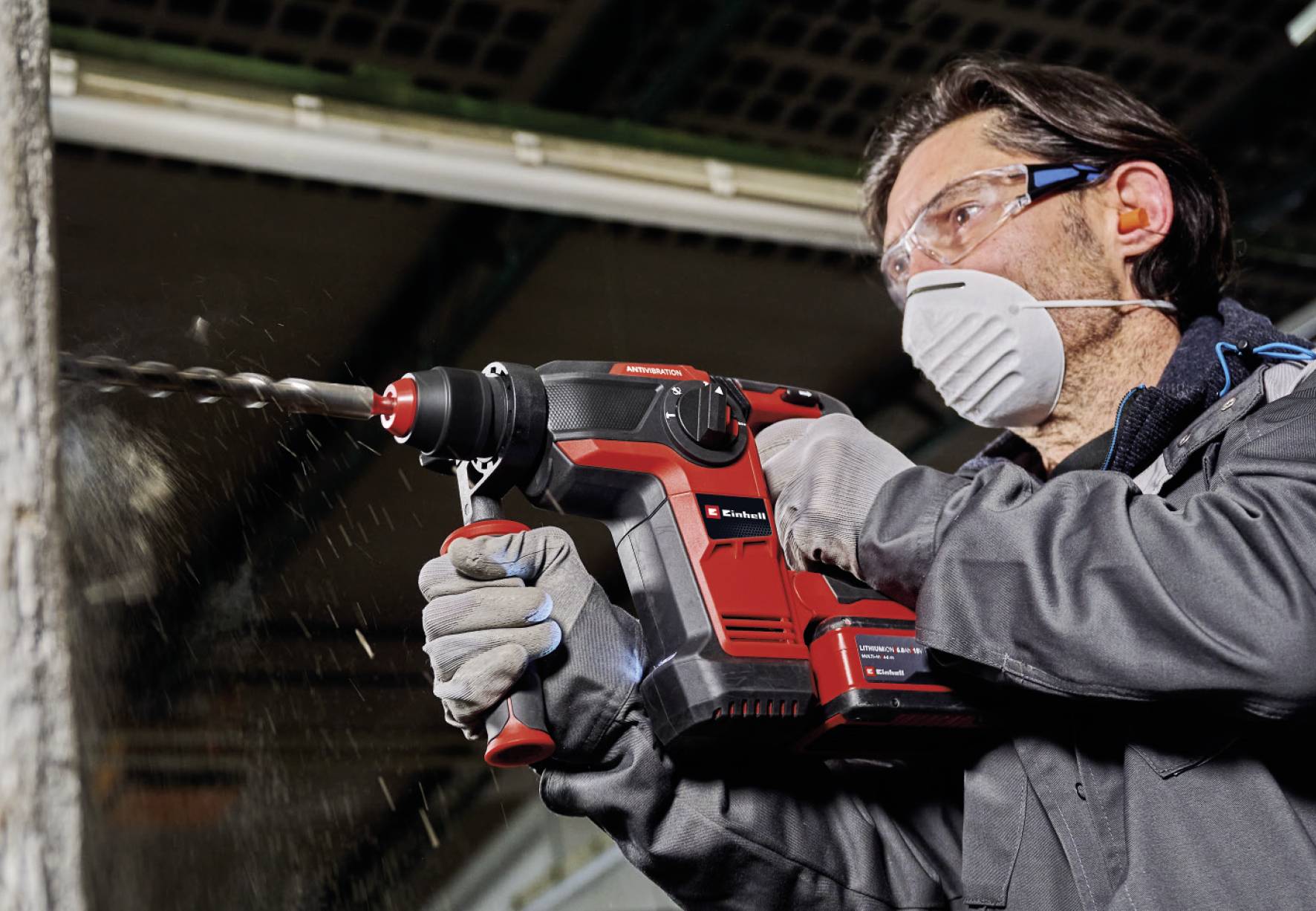 A man in protective clothing is drilling into a wooden wall with a red electric power tool. Working with concentration in a workshop.