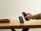 Hand holding smartphone over wireless charging pad on wooden table. Notebook and pen in the background.