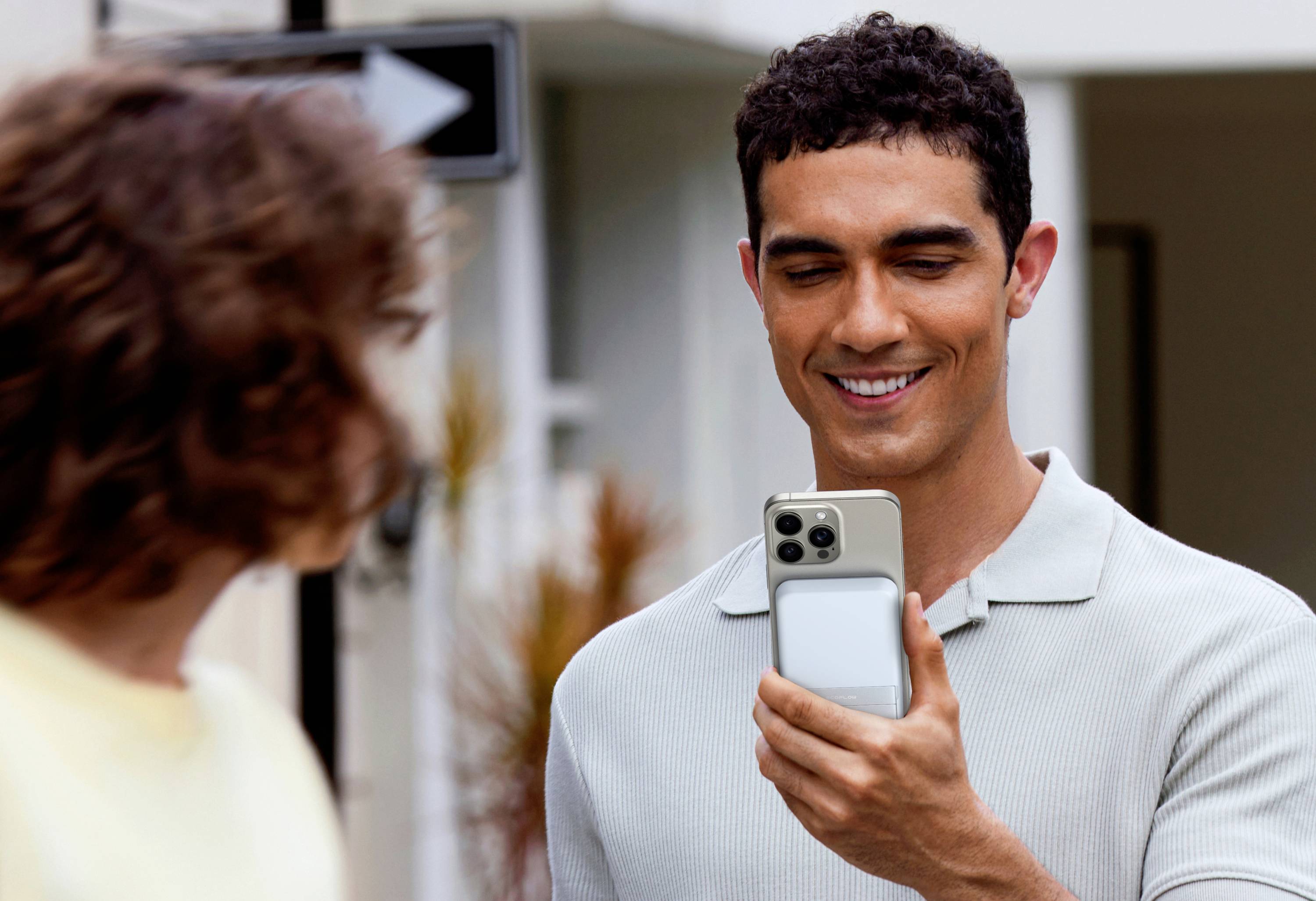 A man is holding a smartphone in his hand, smiling and looking at the screen, with another person blurred in the foreground.