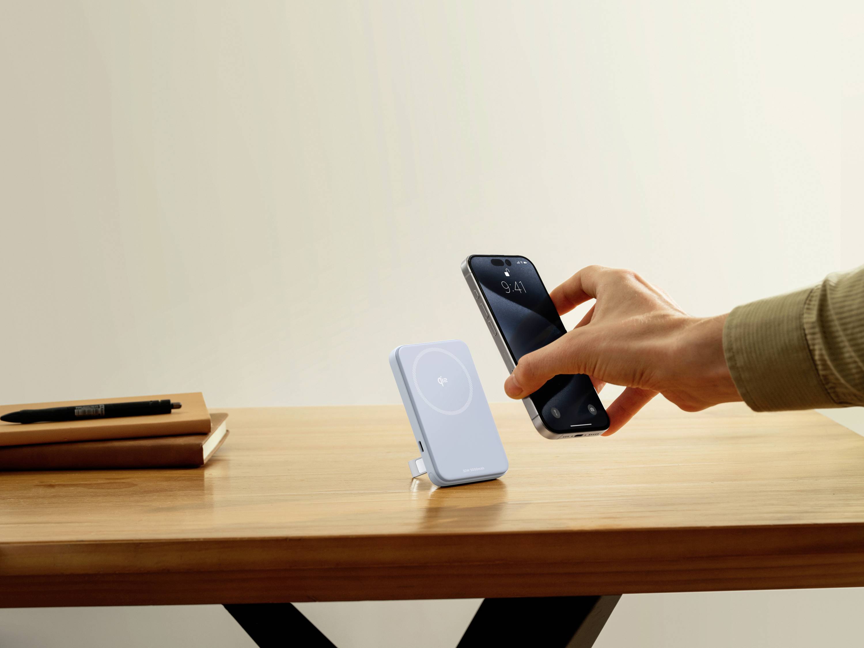 A person holds a smartphone over a wireless charging pad situated on a wooden table. A notebook and a pen are placed beside it.