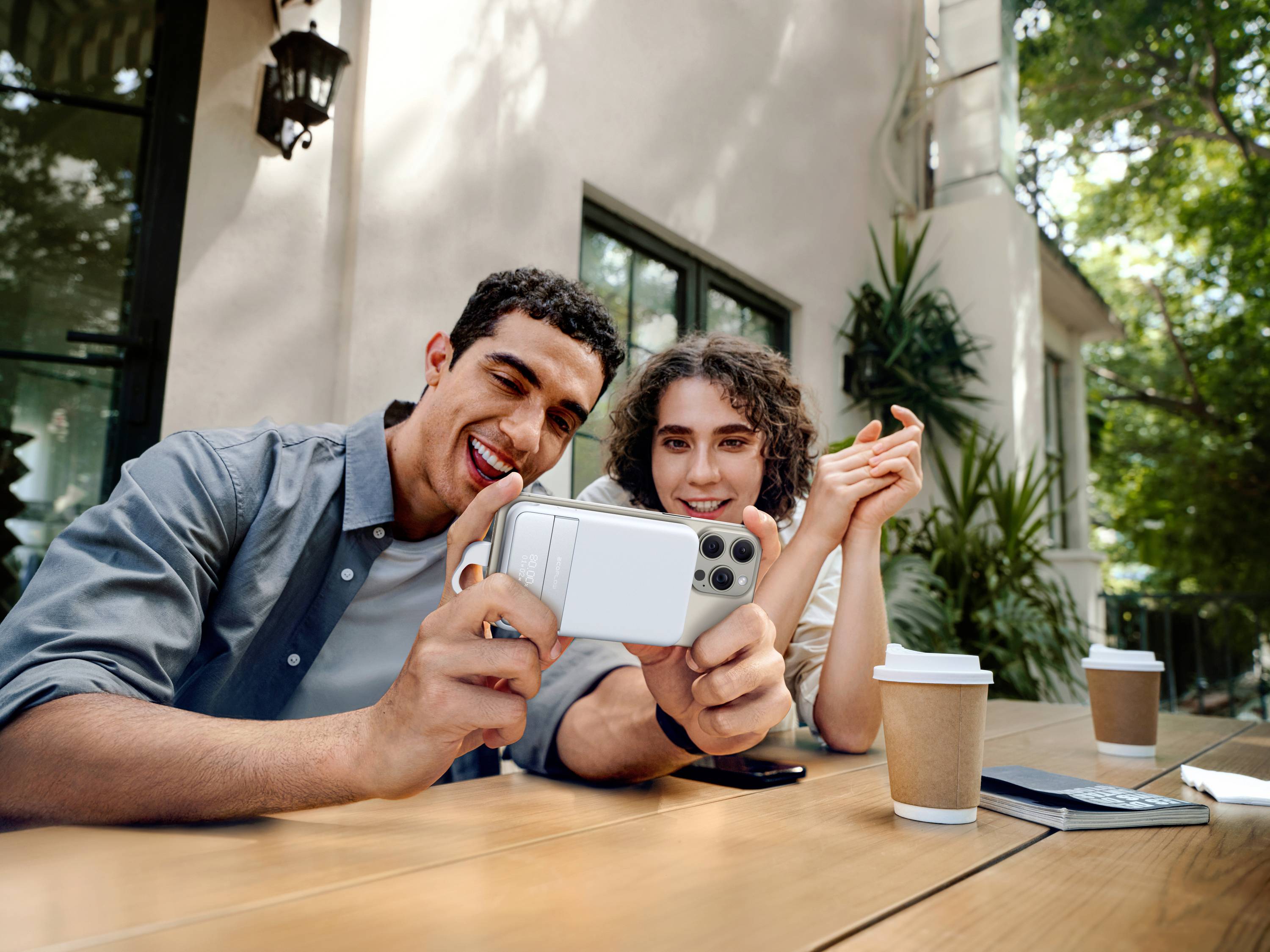 A smiling couple are sitting outside at a table, taking a selfie with a mobile phone. Two coffee mugs are on the table.