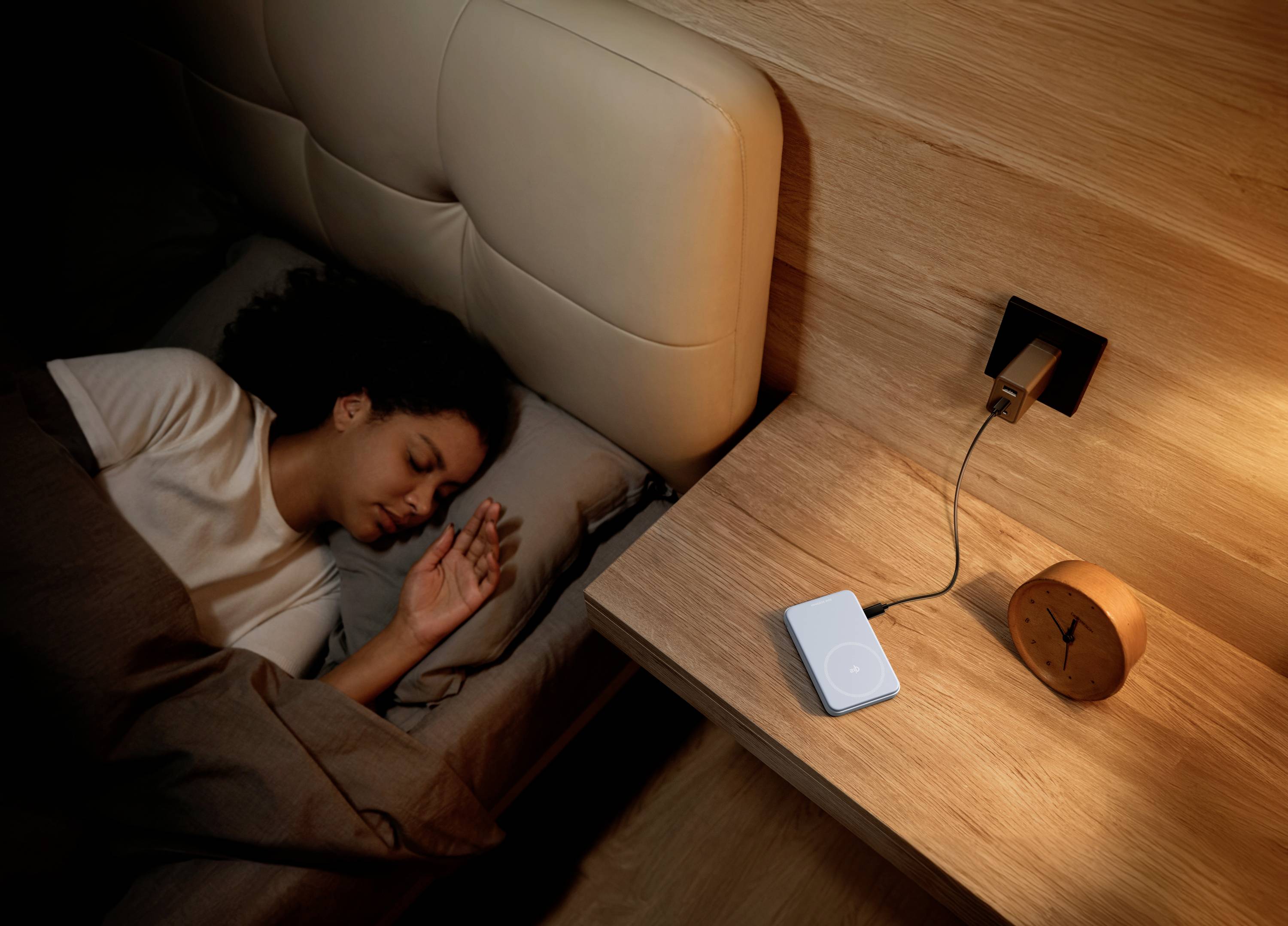 A person is sleeping on a bed in a dark room. Next to it stands a bedside table with a connected clock.