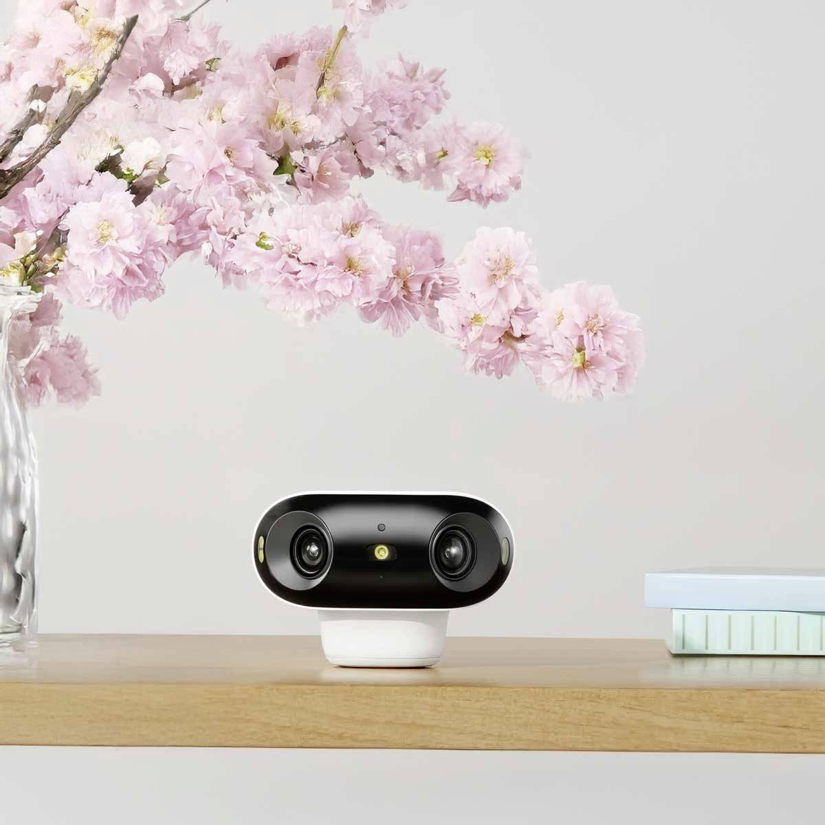 A small, black, spherical camera with two lenses sits on a table next to a glass containing pink cherry blossoms.