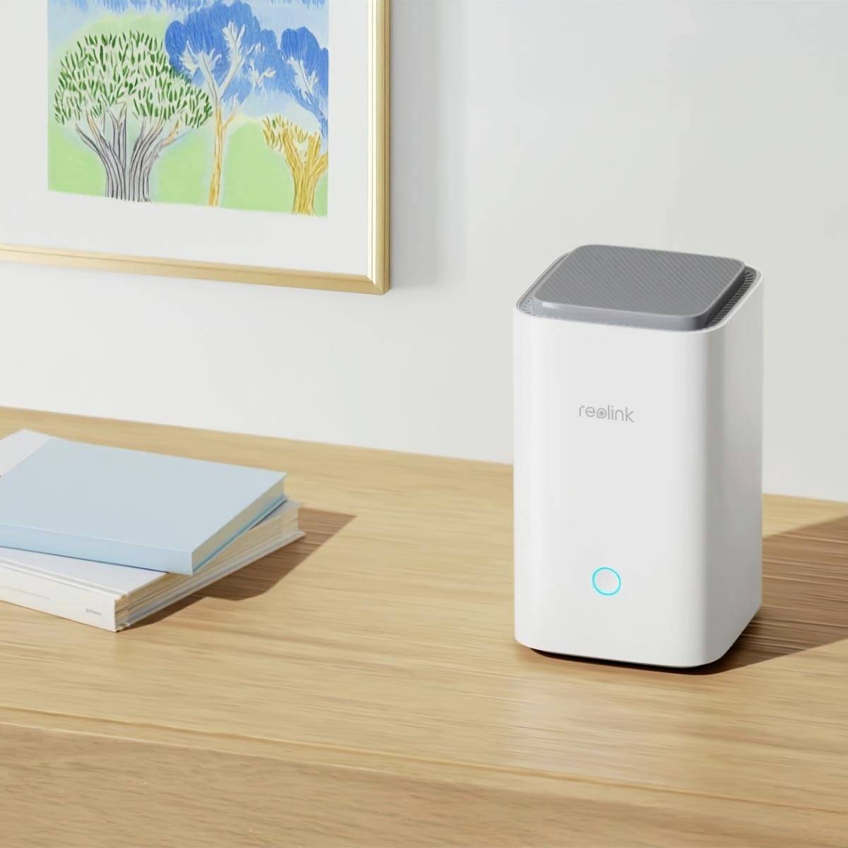 A white Wi-Fi router with an illuminated symbol sits on a wooden table next to a stack of books. A landscape painting hangs on the wall.