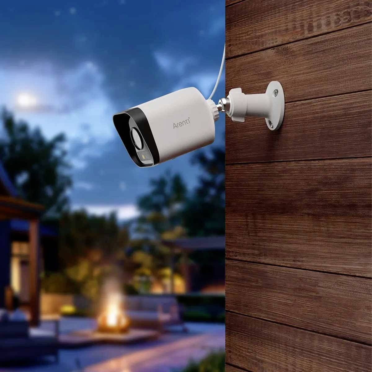 Security camera mounted on a wooden wall, overlooking a garden with a fire pit at dusk.