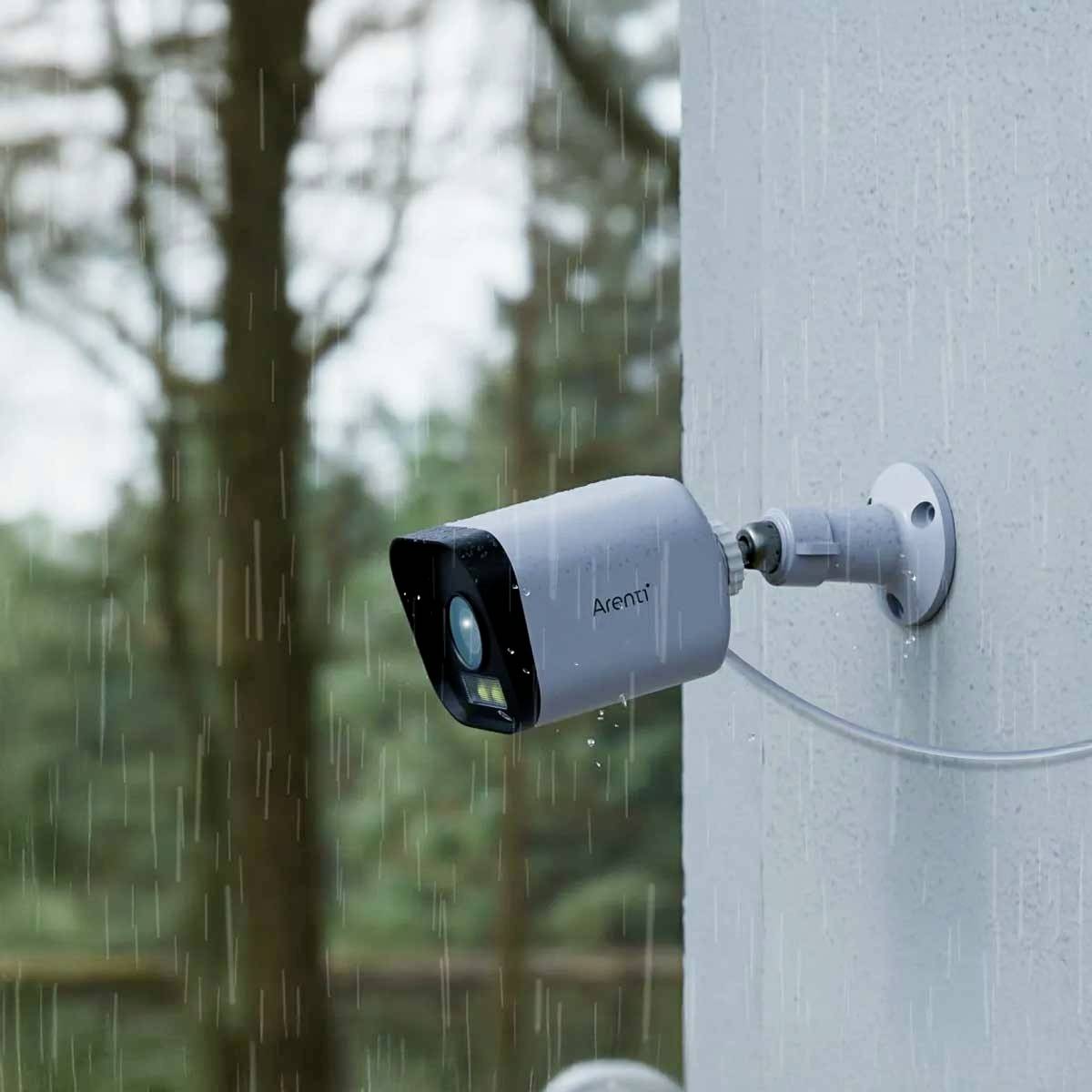A surveillance camera is mounted on a pole. It is raining, and blurred trees can be seen in the background.