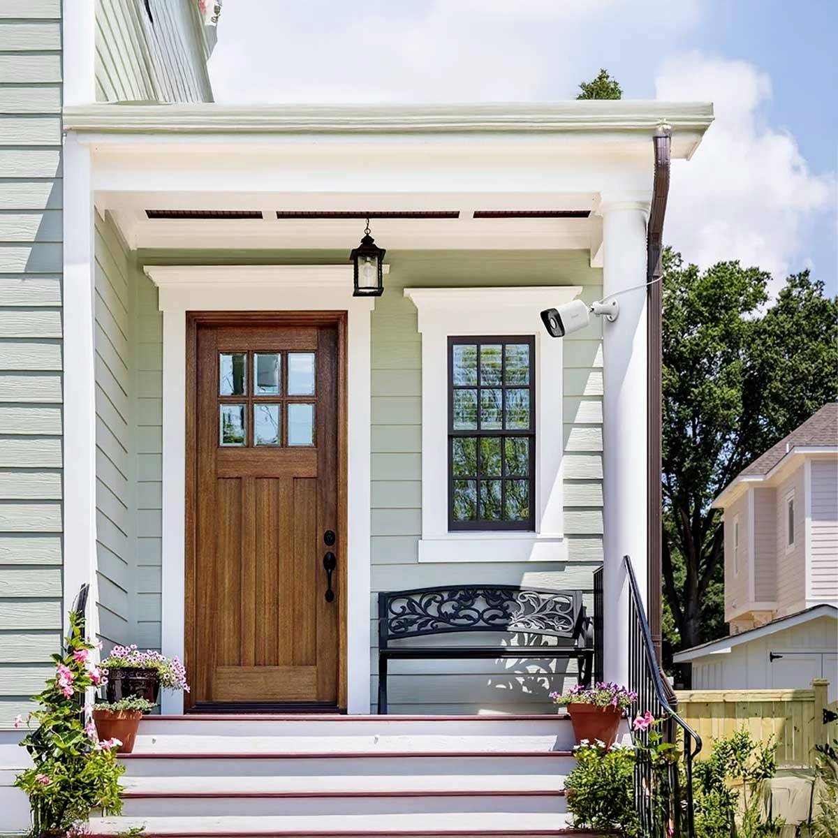 A welcoming entrance area of a house with a light green façade, wooden door, bench, and plants that create a friendly atmosphere.