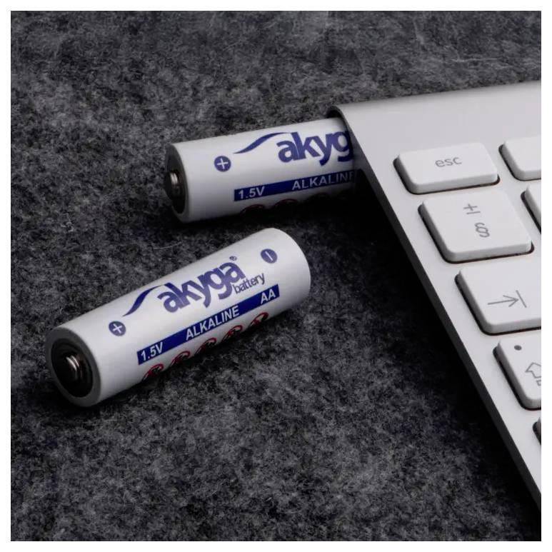 A computer keyboard lies on a grey surface, next to two white AA batteries from the brand 'akyga'.