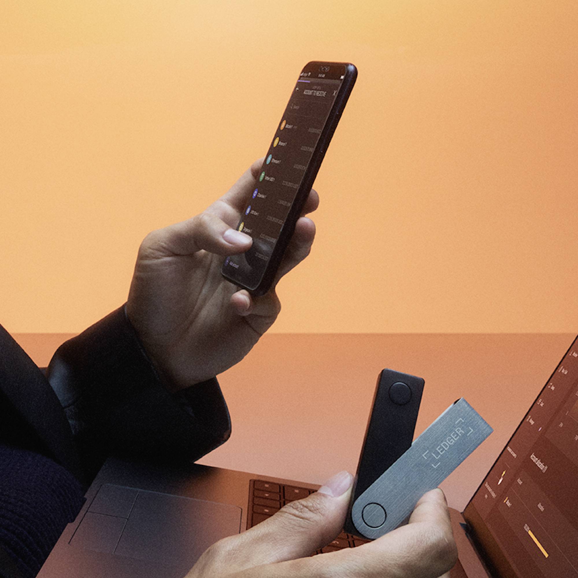 A person is holding a smartphone and a Ledger hardware wallet in front of a laptop. The screens display a cryptocurrency transaction app.