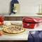 A red pizza maker sits on a kitchen worktop with a freshly baked pizza. Ingredients such as tomatoes and mushrooms are laid out beside it.