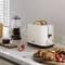 A toaster with browned toast, next to a coffee cup, a jug, bread rolls, and strawberries on a kitchen worktop.