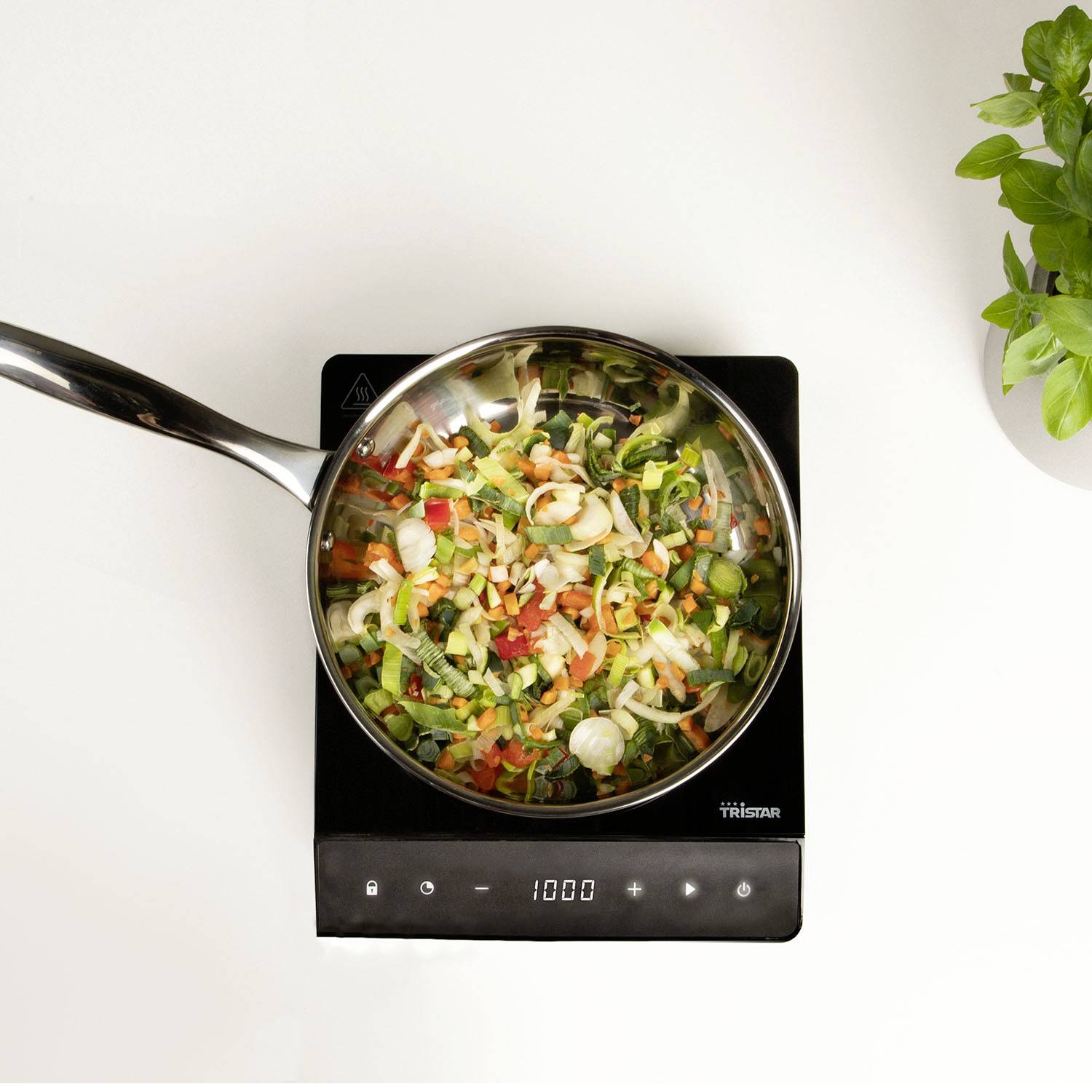 Frying pan with a colourful mix of vegetables on a black induction hob, next to a green basil pot.