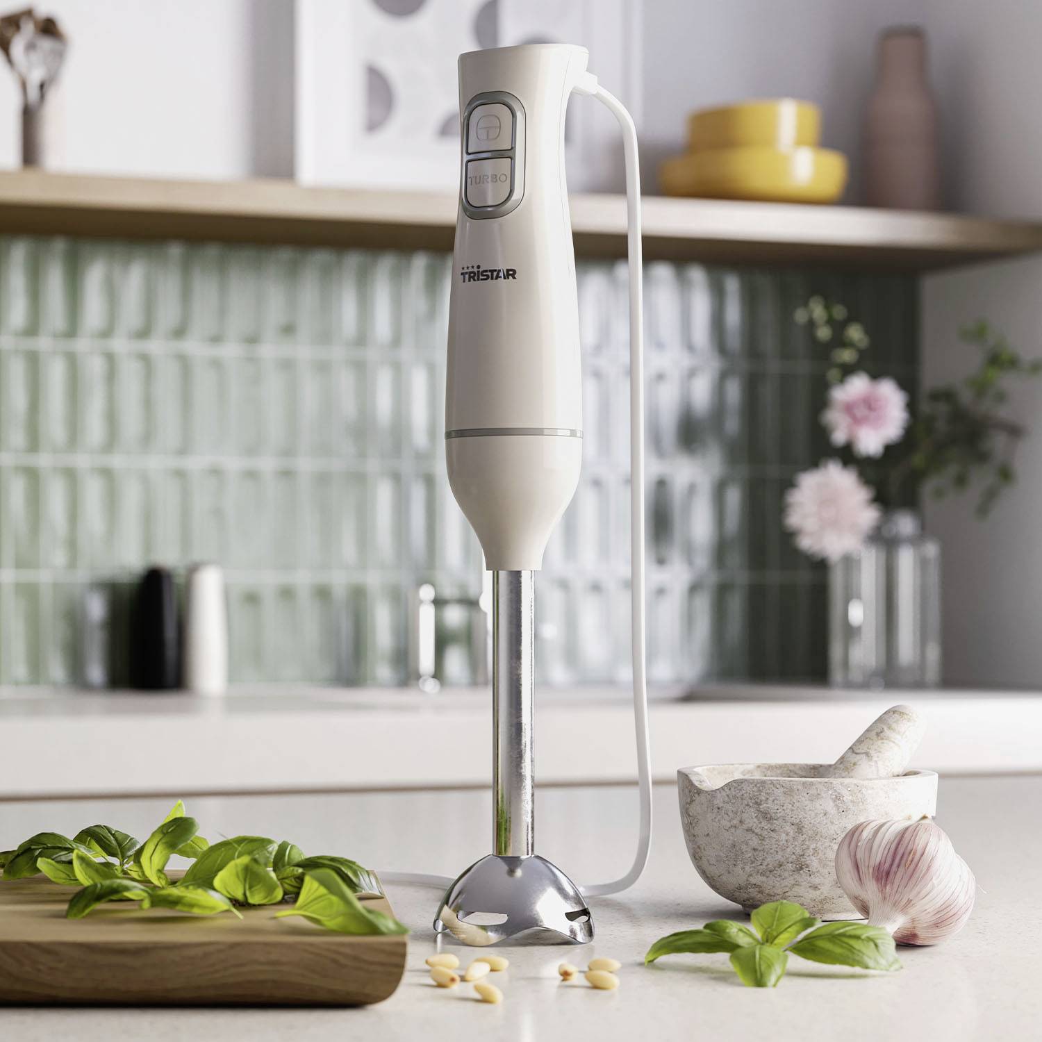A hand blender sits on a kitchen worktop, surrounded by basil, garlic, and a pestle. Tiles are visible in the background.