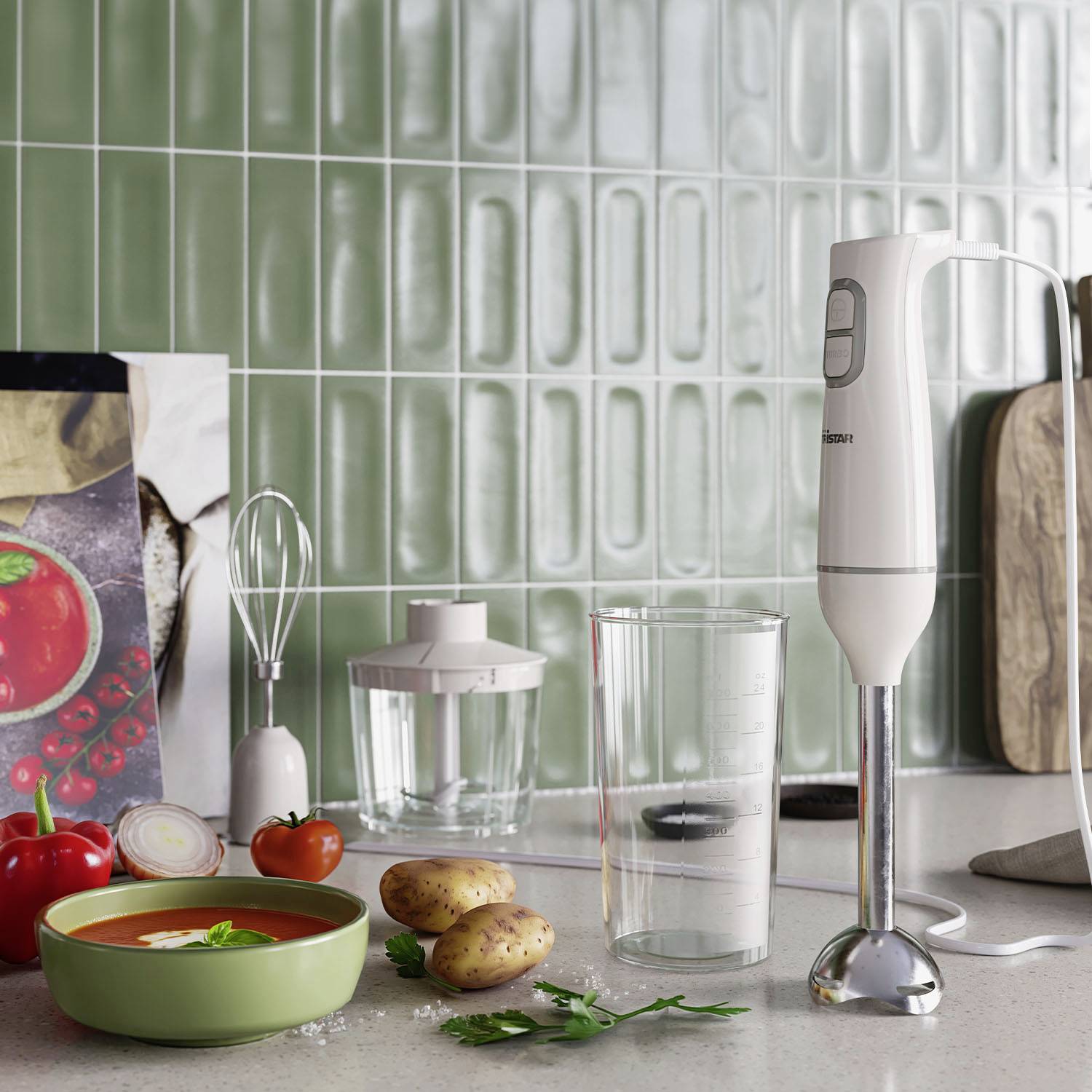 A hand blender on a kitchen worktop, surrounded by a green bowl of soup, three potatoes and a whisk in front of a green tiled wall.