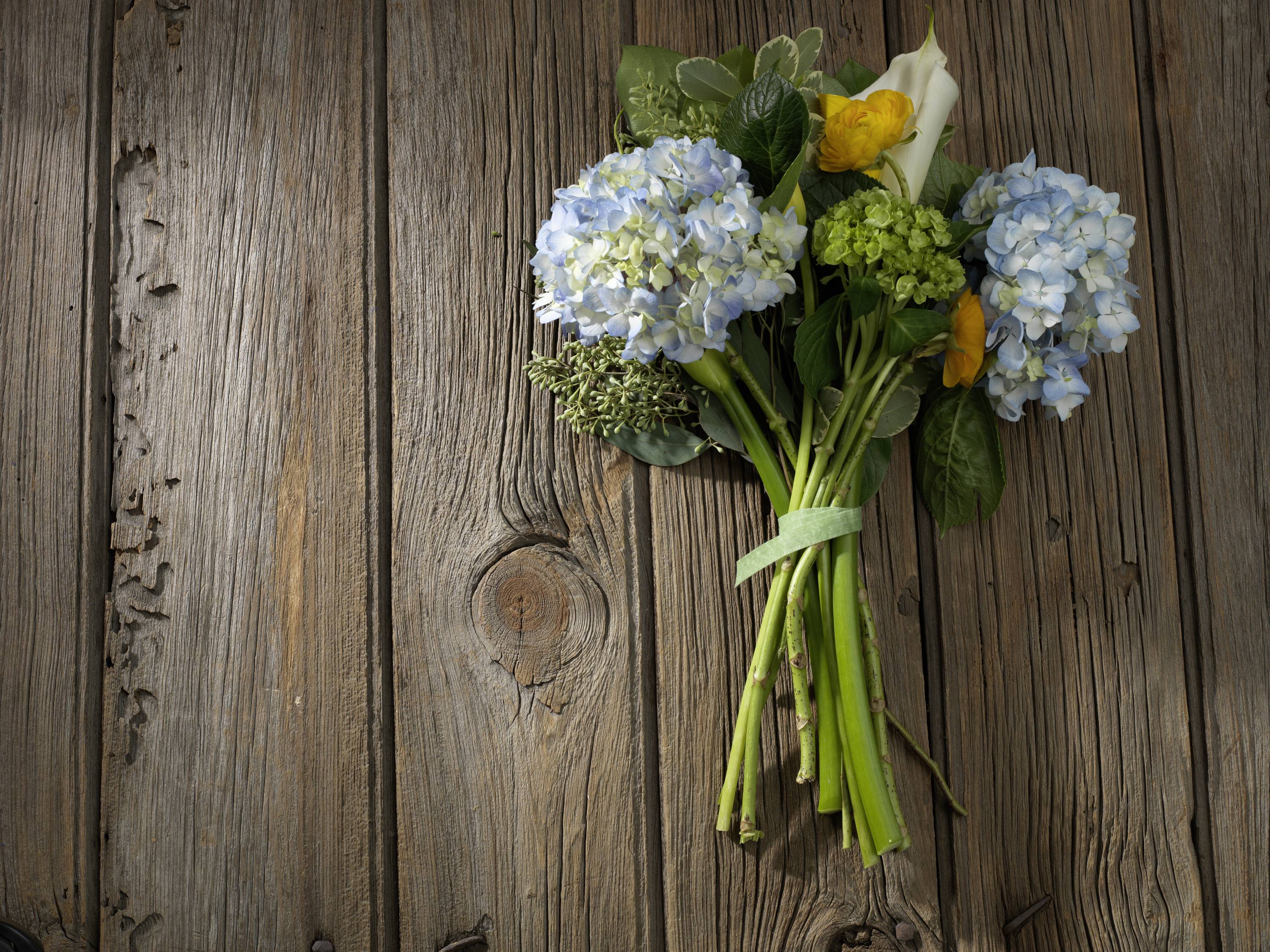 A bouquet of blue hydrangeas, yellow and white flowers rests on a rustic wooden table. Sunlight shadows are visible.