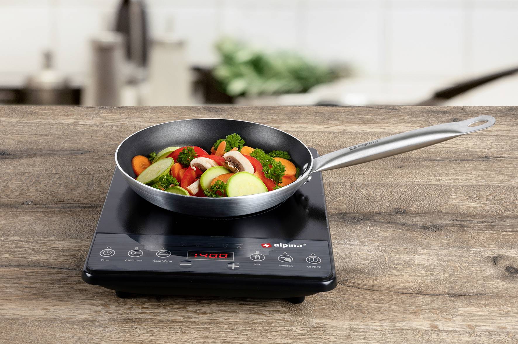 A pan with vegetables is sautéing on an induction hob. It contains broccoli, courgettes and peppers. A kitchen can be seen in the background.