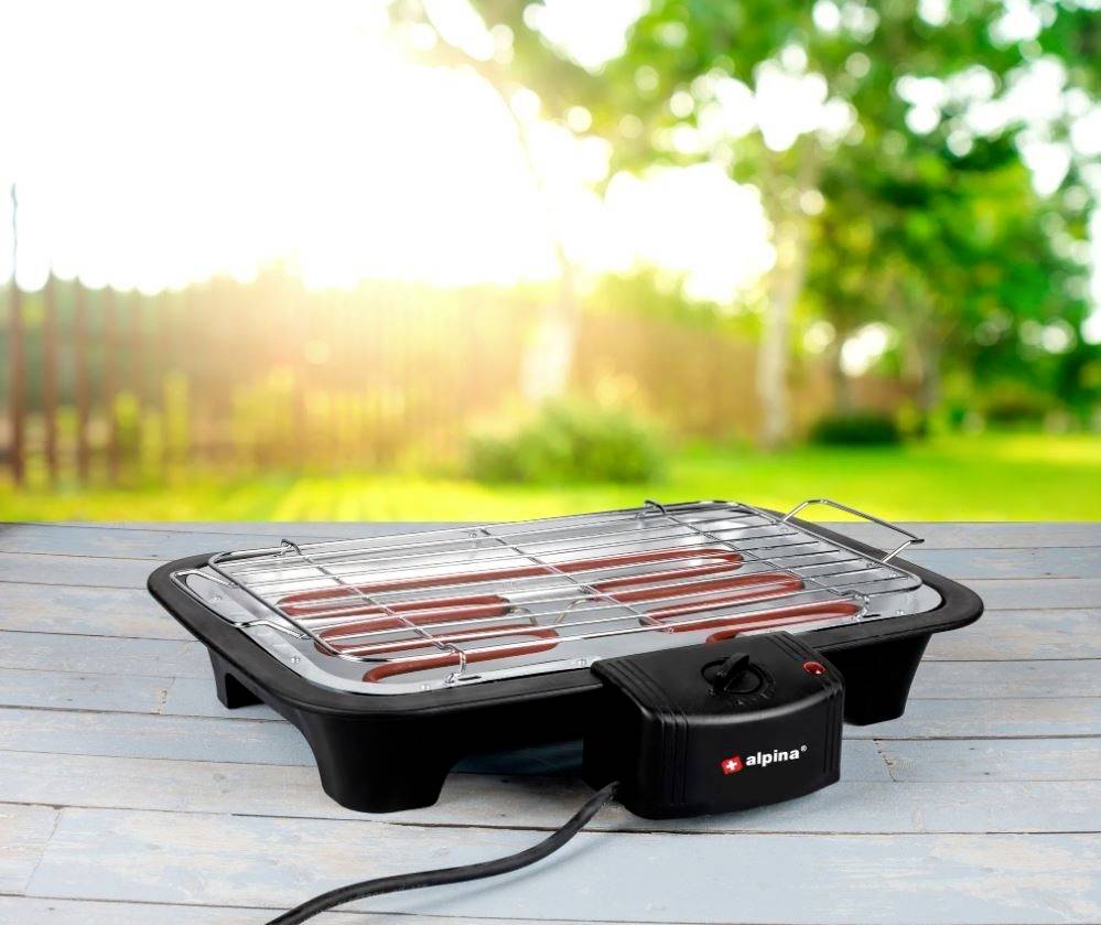 An electric grill with glowing red heating elements stands on a wooden table outdoors. Trees are blurred in the background.